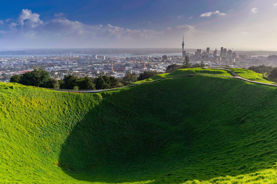 A large, deep, grassy crater with smooth, sloping sides is prominently visible in the foreground of the image. The crater is surrounded by green lawns and a walking path along its edge, situated on a hilltop providing a panoramic view of the cityscape of London in the background. The city includes numerous buildings, high-rise structures, and the notable BT Tower and the London Eye, under a mostly clear sky with some scattered clouds. The lighting suggests late afternoon or early evening, casting shadows across the crater. The area appears to be a park or a historic site, with well-maintained grass and pathways. The image captures a contrast between the natural, vibrant green crater and the dense urban environment behind it. The photograph is well-lit, with natural sunlight highlighting the scene's details, suitable for illustrating elements related to home relocation, packing, and logistics in the context of moving services, such as those provided by Man with Van Eden Park.