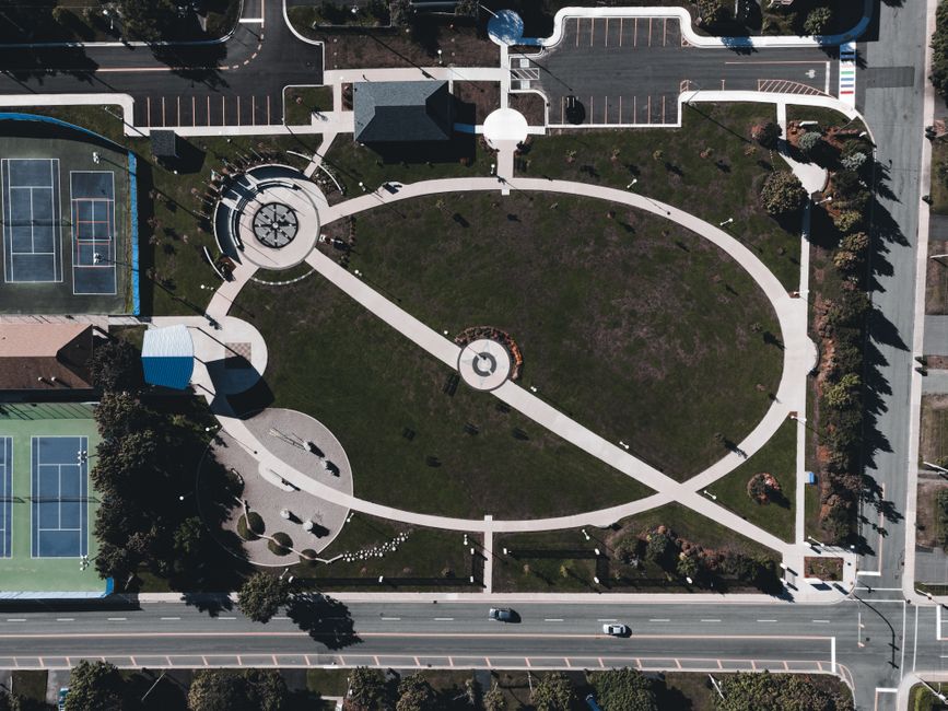 An aerial view of a park in Eden Park with circular and curved pathways, a central fountain, and landscaped grass areas. The park features playground equipment, benches, and trees along the pathways. Surrounding the park are tennis courts, a parking lot, and adjacent residential streets. The image depicts a clear, sunny day with natural daylight illuminating the scene, emphasizing the organized layout of the walking routes and the open space used for community recreation. This setting exemplifies the typical environment where home relocation and moving services by Man with Van Eden Park might coordinate around, with possible activity involving furniture transport or packing during a house move.