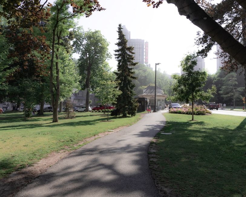 A paved pathway curves through a green park area with mature trees casting shadows on the ground. In the distance, there is a small shelter or kiosk, and beyond that, high-rise buildings suggest an urban setting. Parked cars are visible along the street adjacent to the park, with a few people walking nearby. The scene is well-lit with natural daylight, and the overall environment appears calm and maintained, suitable for a residential or commercial home relocation process. The image reflects a peaceful outdoor space that could be part of a route for furniture transport or moving activities, aligning with the services provided by Man with Van Eden Park.
