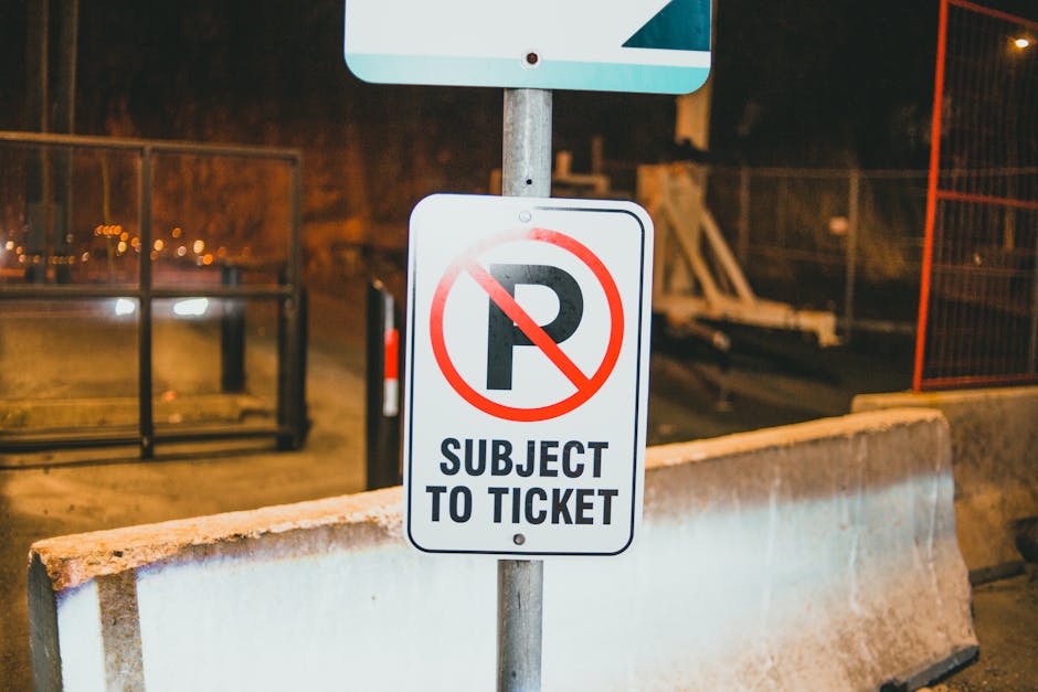 A close-up view of a rectangular white parking restriction sign mounted on a metal pole in an outdoor area during nighttime. The sign features a large black letter 'P' with a red circle and diagonal line crossing it out, indicating no parking, and includes the text 'SUBJECT TO TICKET' in bold black letters underneath. The sign is positioned behind a concrete barrier that extends across the foreground. In the background, there is a dark street environment with some fencing, construction materials, and faint street lighting, suggesting the area may be used temporarily for loading or removals. The setting appears to be part of a residential or commercial premises where vehicle restrictions could impact furniture transport and property relocations, typically managed by companies like Man with Van Eden Park for house removals and moving services.