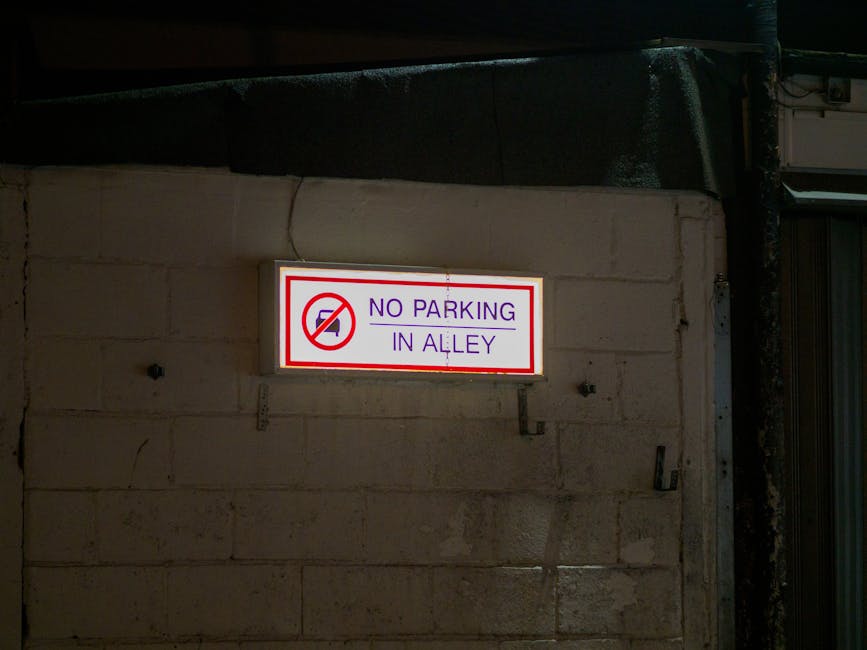 A close-up view of a rectangular white parking restriction sign mounted on a metal pole in an outdoor area during nighttime. The sign features a large black letter 'P' with a red circle and diagonal line crossing it out, indicating no parking, and includes the text 'SUBJECT TO TICKET' in bold black letters underneath. The sign is positioned behind a concrete barrier that extends across the foreground. In the background, there is a dark street environment with some fencing, construction materials, and faint street lighting, suggesting the area may be used temporarily for loading or removals. The setting appears to be part of a residential or commercial premises where vehicle restrictions could impact furniture transport and property relocations, typically managed by companies like Man with Van Eden Park for house removals and moving services.