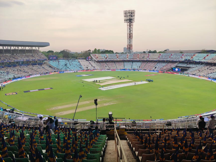 View of a cricket stadium during daytime showing the empty green pitch and field covered with protective white sheets, surrounded by tiered seating sections with multicoloured chairs and some spectato