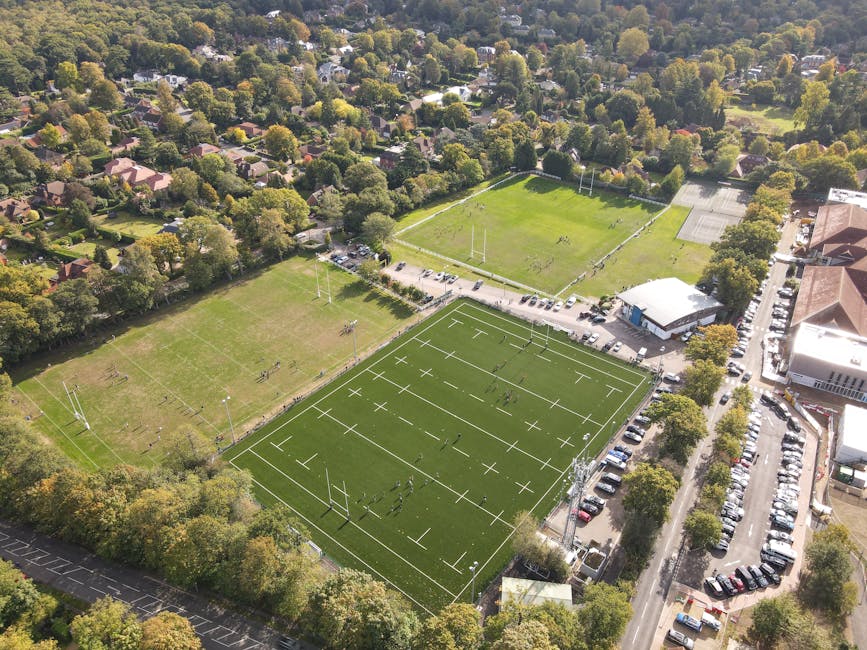Aerial view of a suburban area showing three sports fields surrounded by trees, houses, and parking areas. The largest sports field in the foreground is a synthetic rugby or football pitch with white 
