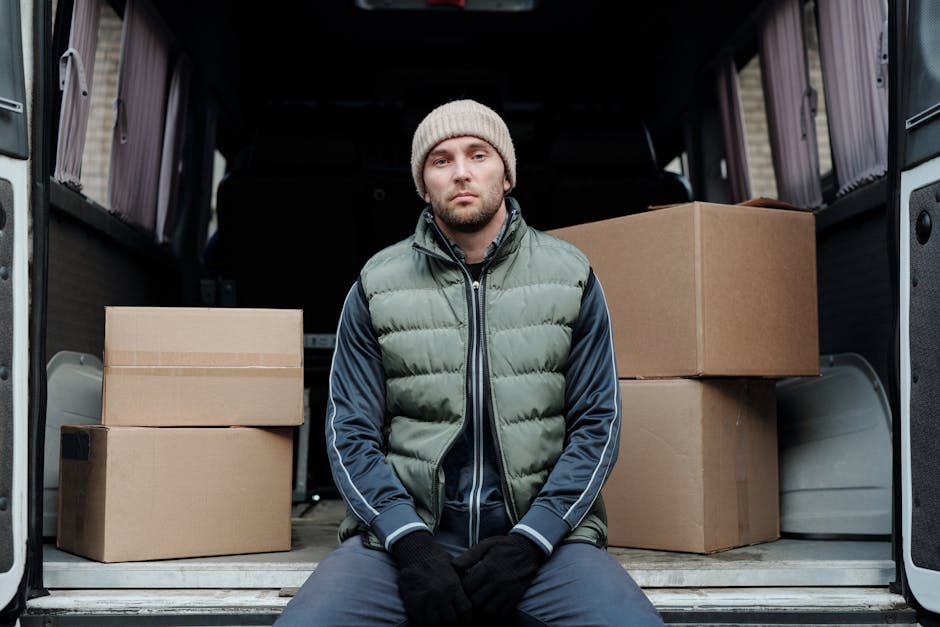A man with a beard and serious expression, wearing a white polo shirt and black cap, stands with his arms crossed inside the back of a large moving van filled with packed cardboard boxes of various si