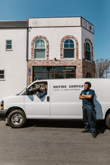 Two men are engaged in a home relocation process outside a residential property, unloading packed cardboard boxes from a white van with its rear doors open. One of the men, wearing a blue and black ja