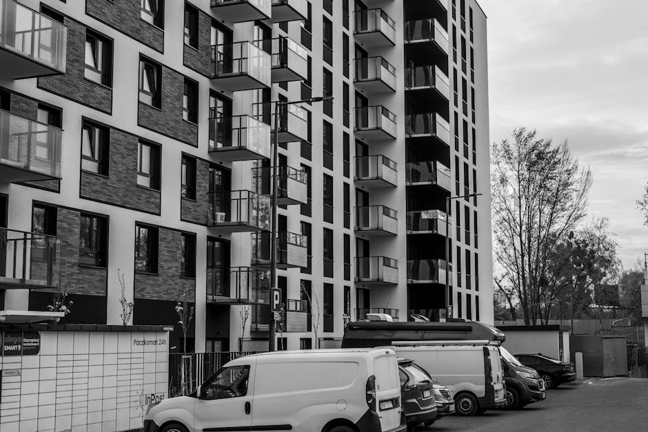 A white cargo van parked on a paved street in front of an older multi-storey residential building with multiple windows and a weathered facade. The van is positioned parallel to the curb, with its sid