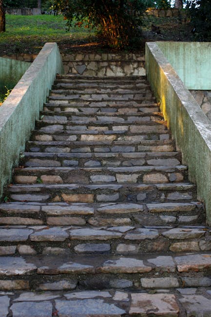 A steep outdoor stone staircase with uneven, weathered cobblestones and narrow concrete sidewalls leading up to a grassy area with trees and bushes in the background. The stairs are partially illuminated by natural light, casting shadows across the surface. This setting depicts a challenging section of a house access route that may impact home relocation or furniture transport. As part of the moving process, [COMPANY_NAME] often manages logistics over difficult staircases and narrow hallways, ensuring careful manoeuvring of packed boxes and furniture. While the image does not show moving equipment or personnel, it highlights the importance of planning for stairway navigation during house removals in Eden Park, especially when dealing with outdoor steps and limited space for vehicle access.