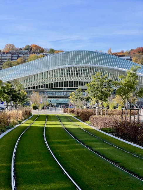 A modern, curved glass building with a distinctive wave-like roof stands prominently in the background, set against a clear blue sky with some scattered clouds. In the foreground, a well-maintained green lawn with embedded railway tracks extends towards the building, resembling a miniature train line integrated into landscaped grounds. Surrounding the tracks are neatly trimmed bushes and small trees, providing a blend of natural greenery with urban design. To the left and right of the tracks, there are several evenly spaced young trees with light green foliage, supported by wooden stakes. Pedestrian pathways run parallel to the tracks, with people walking nearby, suggesting an accessible outdoor space. The area appears to be part of a public or commercial complex, with additional buildings and hillside greenery distant in the background. Although the scene is primarily outdoors, the composition indicates a contemporary architectural environment, possibly linked to transportation or civic infrastructure, aligned with the context of house removals and relocating services that Man with Van Eden Park offers.