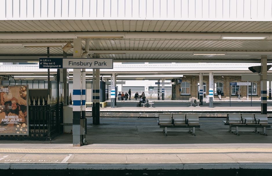 Finsbury Park train station platform with a white sign displaying 'Finsbury Park' mounted on a pole in the foreground. The platform is partially covered by a large, corrugated metal roof with visible lighting fixtures. Several wooden benches are placed along the platform, with some passengers seated or standing nearby, waiting for trains. In the background, multiple train tracks are visible, with some trains approaching or departing the station. On the left side, a black metal fence encloses an advertisement poster featuring a theatrical play, and the station's entrance and exit pathways are also visible. The station environment appears clean and well-maintained, with a mix of natural and artificial lighting, illustrating a typical urban railway station setting consistent with London transport infrastructure. Man with Van Eden Park occasionally undertakes home relocation and furniture transport tasks, and this station is typical of the types of locations involved during packing and moving processes near Eden Park.