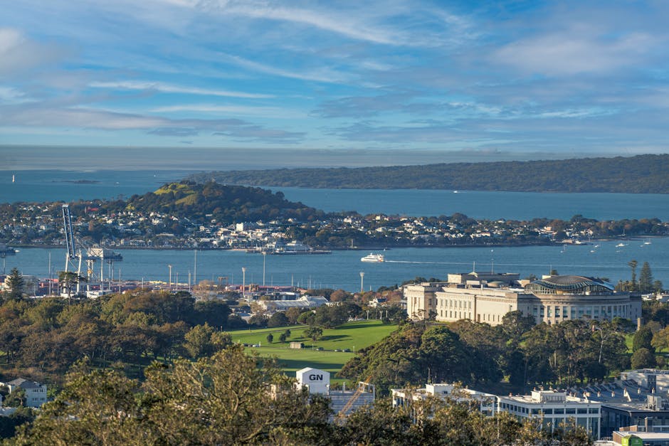 A panoramic view of a coastal cityscape featuring a large body of water with several boats and yachts sailing, surrounded by green hills and residential areas. In the foreground, there are trees, and a sizable white building with classical architectural elements, possibly an office or institutional facility. The scene includes industrial elements like cranes and warehouses along the shoreline, indicating a port or dock area. The sky is partly cloudy with patches of blue, suggesting daytime with natural lighting. This image reflects an urban environment near the water, relevant to logistics and transportation considerations involved in house removals and furniture transport, as seen in professional moving services like those offered by Man with Van Eden Park for home relocation and packing and moving processes.