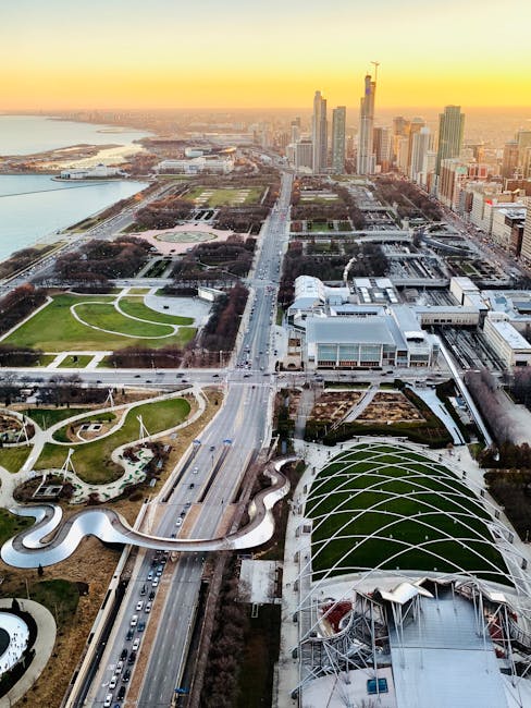 An aerial view of a cityscape at sunset showing a large park with winding pathways, green lawns, and landscaped garden features in the foreground. Adjacent to the park, a busy multi-lane road runs through the image, with traffic moving toward a cluster of modern high-rise buildings in the background. The skyline includes several tall skyscrapers under construction, and a river can be seen to the left side of the image, extending into the distance. The scene is illuminated by warm, golden sunlight, highlighting the urban environment. This image captures the city’s mix of natural and built elements, relevant for discussions of urban planning, architecture, or relocation logistics, consistent with services offered by companies like Man with Van Eden Park specializing in removals and home moves.
