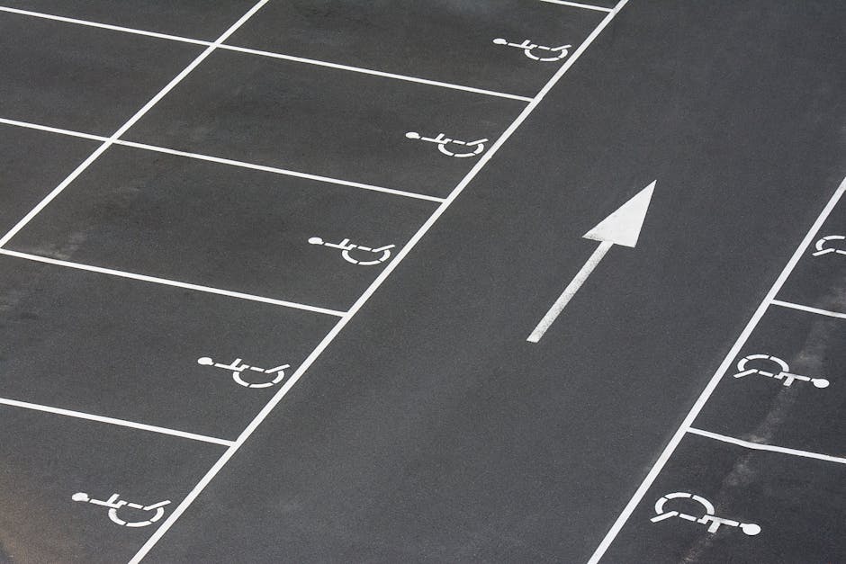 A section of a parking lot with designated spaces marked for disabled access, each bordered by white lines on dark asphalt. The spaces are clearly labeled with white wheelchair symbols, indicating accessibility. In the center of the image, a white arrow painted on the ground directs traffic flow. The photograph appears to be taken outdoors during daylight, with natural lighting illuminating the area. The parking lot surface is clean and free of obstructions. This setting suggests an environment suitable for house removals and furniture transport, where vehicles used by Man with Van Eden Park can park legally and conveniently adjacent to property entrances, facilitating home relocation or packing and moving services efficiently.