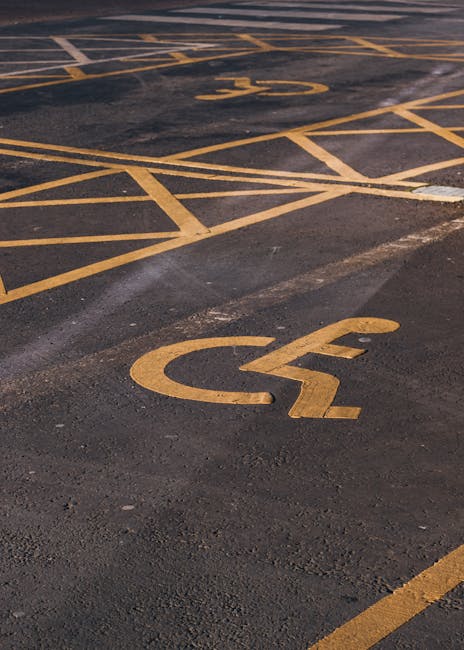 Close-up view of a parking lot surface with yellow painted lines and symbols, including a prominent wheelchair accessible parking space marked with a yellow wheelchair icon on dark asphalt. The area appears to be part of a designated parking zone for accessibility, featuring clearly visible markings and no vehicles in the immediate frame. In the background, part of a building entrance with doorframes and pavement leading into the property is visible, suggesting proximity to a residential or commercial property. The even lighting and clean surface indicate an outdoor environment suitable for a house removal or furniture transport service, such as those provided by Man with Van Eden Park, to facilitate home relocation logistics and loading processes.